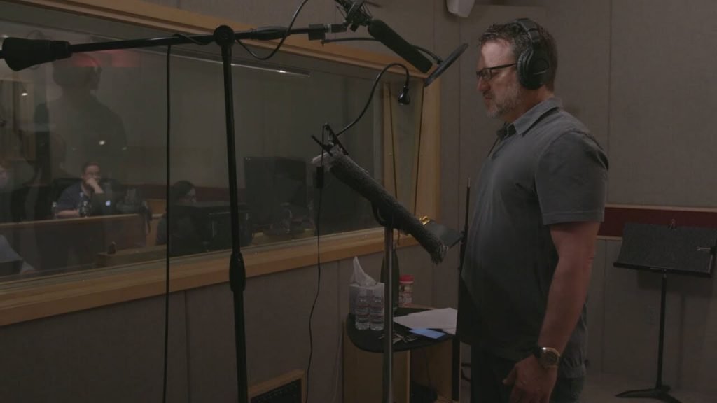 Picture of Steve Blum at the Mic in front of a wide booth window with several people at computers on the other side